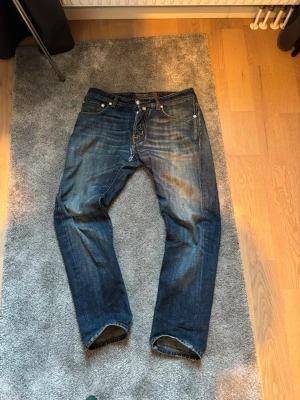Jacob Cohen - Säljer ett par klassiska blå jeans från Jacob Cohen med snygg slitning.  Jeansen har orangea sömmar och en läderpatch bak i midjan. Perfekta för en avslappnad och soft stil. Den sitter slim/rakt. 