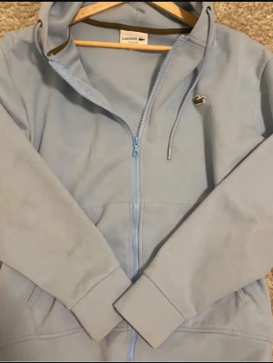 Ljusblå hoodie från Lacoste - Snygg ljusblå hoodie från Lacoste med dragkedja framtill och klassisk krokodil-logga på bröstet. Hoodien har huva med dragsko och två stora fickor framtill. Tillverkad i mjukt material som känns skönt mot huden. Perfekt för en avslappnad och stilren look.