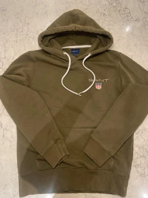 Olivgrön hoodie från GANT - Snygg olivgrön hoodie från GANT med vit dragsko i huvan och broderad logga samt sköld på bröstet. Klassisk känguruficka framtill och ribbade muddar vid ärmslut och nederkant. Perfekt för en avslappnad och stilren look.