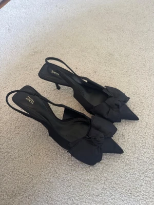 Svarta pumps med rosett från Zara - Säljer ett par eleganta svarta pumps från Zara med spetsig tå och låg klack. Skorna har en snygg rosettdetalj framtill och öppen häl med rem. Tillverkade i textilmaterial som ger en lyxig känsla. Perfekta för dig som vill ha en chic och trendig look. De är i storlek 37, men jag anser att de även passar storlek 38 bra! 💞 Jag har storlek 38,5 och de är bara lite lite försmå för mig!