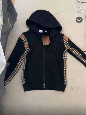  Burberry zip - Snygg svart hoodie från Burberry med dragkedja och klassiska beige-rutiga paneler längs ärmarna och axlarna. Huva med dragsko och fickor framtill. Perfekt för dig som vill ha en exklusiv streetwear-look. Materialet känns mjukt och bekvämt.