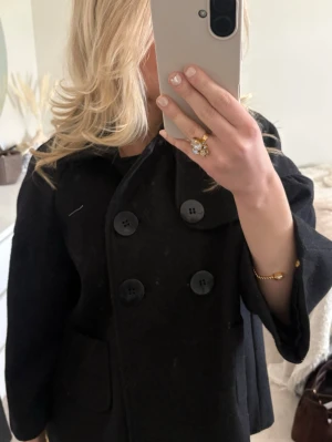 Svart kappa från Gianni - Stilren svart ullrock från Gianni med dubbelknäppning och stora knappar framtill. Rocken har bred krage, två framfickor och en rak, något kortare passform. Perfekt för dig som gillar klassisk och enkel stil.