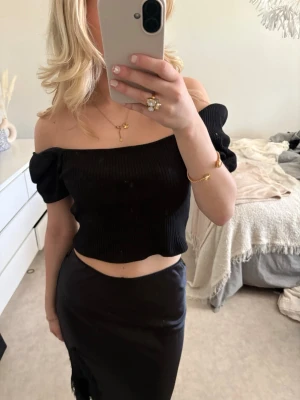 Svart croppad offshoulder topp - Trendig svart croppad topp med ribbad struktur och puffiga offshoulder-ärmar. Toppen har en smickrande passform och är perfekt för dig som vill ha en stilren men ändå edgy look. Materialet känns mjukt och stretchigt, vilket gör den bekväm att bära.