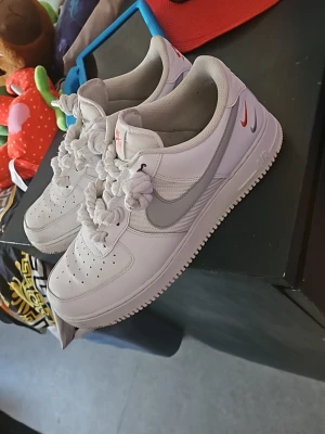 Nike vita Air Force 1 sneakers - Klassiska vita Nike Air Force 1 sneakers med grå swoosh och röda detaljer på hälen. Skorna har perforerad tå, tjock vit sula och unika snörningar med knutar. Tillverkade i läder och textil för en clean och tidlös look.