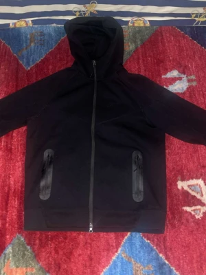 Nike tech  - Säljer en svart hoodie från Nike med hel dragkedja och två fickor med dragkedja på framsidan. Hoodien har en huva och Nike-loggan i silver på bröstet. Materialet är mjukt och stretchigt, perfekt för en sportig och avslappnad look.