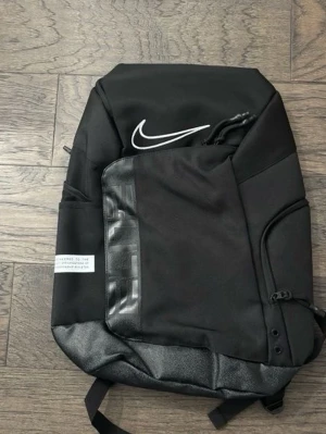 Svart ryggsäck från Nike - Snygg svart ryggsäck från Nike med vit logga på framsidan. Ryggsäcken har flera fack, dragkedja och detaljer i både tyg och konstläder. Perfekt för skolan eller träningen och har en modern, stilren design.