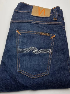 Nudie Jeans Lean Dean blå jeans - Säljer ett par klassiska blå jeans från Nudie Jeans, modellen Lean Dean. Jeansen har orangea sömmar, vit brodyr på bakfickan och är gjorda i 98% bomull. Snygg passform och tidlös design som funkar till allt. Jeansen är inte använda. Kan tänka mig att gå ner i pris vid snabb affär.