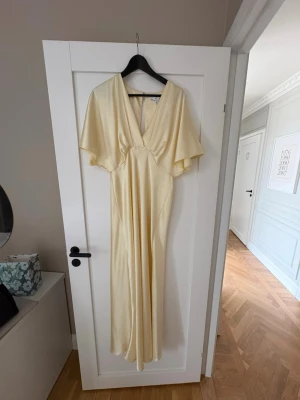 Ljusgul långklänning med v-ringning - Elegant ljusgul långklänning med djup v-ringning och vida, korta ärmar. Klänningen har ett mjukt, glansigt material som ger ett exklusivt intryck och ett flytande fall. Perfekt för sommarens speciella tillfällen.