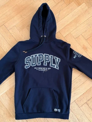 Svart hoodie med tryck SUPPLY - Svart hoodie med stor vit SUPPLY-text på bröstet och tryck på ena ärmen. Klassisk känguruficka framtill och justerbar huva med snören. Mjuk bomullsblandning, perfekt för en avslappnad streetwear-look.