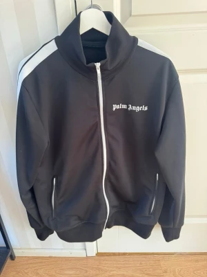 Palm Angels svart track jacket XL - Svart track jacket från Palm Angels med vit dragkedja och vita ränder på axlarna. Tröjan har hög krage, två fickor med dragkedja och logga på bröstet. Tillverkad i 100% polyester för en sportig och modern look.  NECKTAG finns inte 