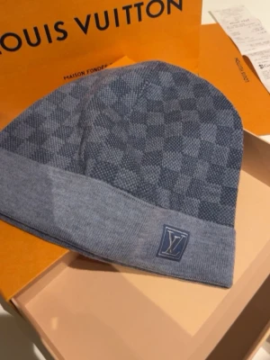 Blå Louis Vuitton mössa med rutmönster - Snygg blå mössa från Louis Vuitton med klassiskt rutmönster och vikt kant. LV-logga framtill och diskret etikett på insidan. Perfekt accessoar för dig som vill ha något exklusivt och stilrent. Stickad i mjukt material för extra komfort.