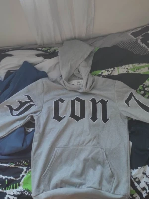 Grå hoodie från Just Man med tryck - Snygg grå hoodie från Just Man med stor svart text 'icon' på bröstet och coola tribal-liknande tryck på ärmarna. Mjuk insida och klassisk känguruficka framtill. Perfekt för en avslappnad streetstyle-look.