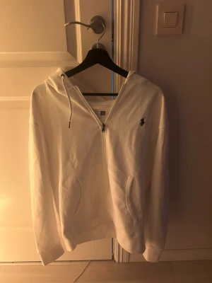 Ralph Lauren zip hoodie - Är 183 på andra bilden, Vit hoodie med dragkedja från Polo Ralph Lauren. Klassisk design med huva och dragsko, två fickor framtill och broderad logga på bröstet. Perfekt för en clean och stilren look. Mjuk bomullskänsla och relaxed fit.