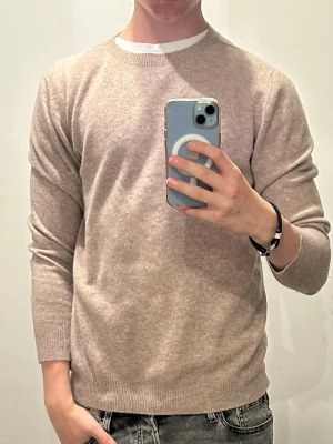 Beige rundhalsad cashmeretröja - Säljer en stilren beige tröja i mjuk cashmere med rund halsringning och långa ärmar. Tröjan har ribbade muddar vid ärmslut och nederkant, perfekt för dig som gillar en enkel men snygg look. Passar till jeans eller chinos.