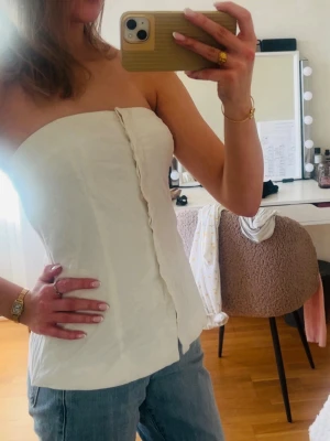 Vit axelbandslös tubtopp - Säljer en stilren vit tubtopp i linnematerial. Toppen är axelbandslös och har en rak, bandeau-liknande form med en liten slits framtill. Perfekt att matcha med jeans eller kjol för en fräsch look.