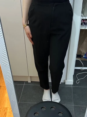 Svarta kostymbyxor med avsmalnande ben - Snygga svarta kostymbyxor med hög midja och avsmalnande ben. Byxorna har fickor i sidorna och stängs med dragkedja och knapp framtill. Perfekta för en stilren look och passar till många olika outfits.