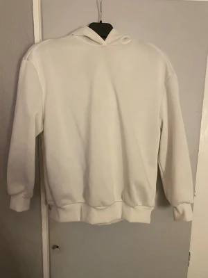 Vit hoodie med tryck och huva - Säljer en vit hoodie med stor svartvit print på ryggen som visar två händer med smycken och texten 'Rock the world'. Hoodien har huva, ribbade muddar och är gjord i mjukt material. Perfekt för dig som gillar streetstyle och vill sticka ut.