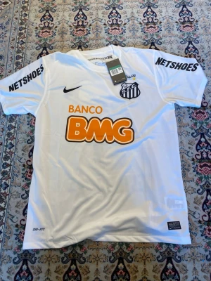 Vit Santos FC matchtröja Nike - Säljer en vit Santos FC matchtröja från med tryck 'BANCO BMG' och 'NETSHOES' på ärmarna. Tröjan har klubbens emblem på bröstet, nummer 11 och Neymar JR på ryggen. Materialet är Dri-FIT polyester som andas och passar perfekt för fotboll.