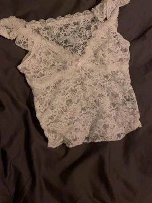 Vit lace tröja med breda axelband - Säljer en vit lace tröja i genomskinlig spets med breda axelband och vacker blommig struktur. Perfekt för dig som gillar romantiska detaljer och ett feminint uttryck. Materialet är mjukt och följsamt, och spetsen har en fin scallop-kant längs axelbanden.