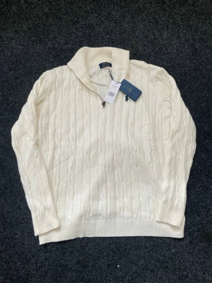 Vit kabelstickad tröja från Ralph Lauren - Säljer en vit kabelstickad tröja från Ralph Lauren med halv dragkedja och ribbad krage. Tröjan har ribbade muddar och är tillverkad i mjukt material. Perfekt för dig som vill ha en klassisk och stilren look. tröjan sitter som en XL, men står 2XL