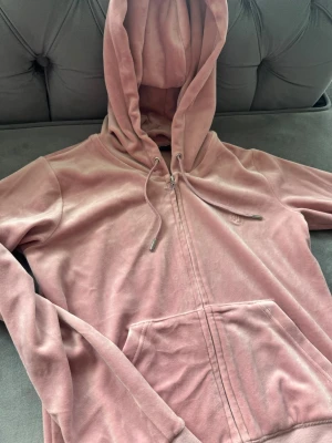 Rosa velour hoodie med dragkedja - Mjuk rosa hoodie i velour med dragkedja framtill och stor ficka. Huva med snören och ribbade muddar. Perfekt för chill dagar och har en diskret logga på bröstet. Snygg och bekväm att bära när du vill ha en avslappnad look.