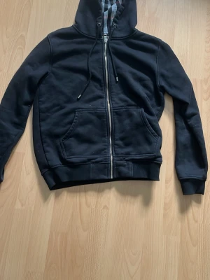 Svart hoodie från Burberry - Svart hoodie med dragkedja från Burberry, har klassiskt rutigt foder i huvan och mjukt fleecefoder på insidan. Utrustad med två stora fickor framtill och ribbade muddar. Perfekt för dig som vill ha en stilren och bekväm hoodie. STORLEK M men passar S också