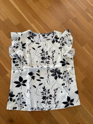 Topp med blommigtmönster  - Härlig topp med ett vackert svartvitt blommönster och söta volangärmar. Lätt och luftig modell som passar perfekt till våren och sommaren.