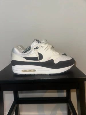 Nike Air Max 1 vita och svarta sneakers - Nike Air Max 1 sneakers i vitt och svart med klassisk synlig Air-enhet i sulan. Ovandelen är i mesh och läder med svarta detaljer och Swoosh-logga på sidan. Lågt skaft och broderad Nike Air-logga på hälen. Perfekta för dig som gillar retrostil.