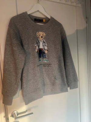 Grå sweatshirt Polo Bear Ralph Lauren - Helt ny Grå sweatshirt från Ralph Lauren med ikoniskt Polo Bear-tryck framtill. Tröjan har rund halsringning, ribbade muddar och avslappnad passform. Perfekt för en chill och trendig look. Materialet är mjukt och bekvämt, troligen bomullsblandning.
