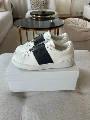 Vit sneaker med svart band från Valentino - Snygga vita sneakers från Valentino med ett brett svart band över sidan och coola nitar på hälen. Skorna är gjorda i läder och har en klassisk låg profil med vita snören och mönstrad sula. Perfekt för dig som vill ha en stilren och trendig look.