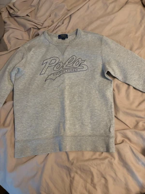 Grå sweatshirt från Polo Ralph Lauren - Snygg grå sweatshirt från Polo Ralph Lauren med broderad logga framtill. Tröjan har rund halsringning, långa ärmar och ribbade muddar. Perfekt för en avslappnad och stilren look.