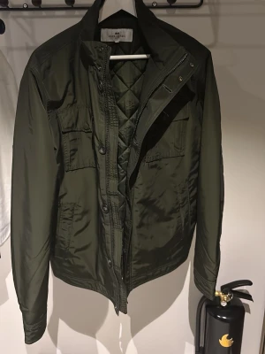 Mörkgrön vindjacka från John Henric - Stilren mörkgrön vindjacka från John Henric med quiltat innerfoder, hög krage och flera fickor framtill. Jackan har dragkedja och knappar, samt justerbara detaljer i midjan. Perfekt för blåsiga dagar 