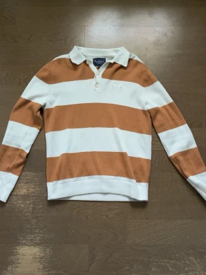 Randig rugbytröja från Bondelid - Säljer en snygg rugbytröja från bondelid med breda horisontella ränder i vitt och rostbrunt. Tröjan har krage, knäppning med två knappar och broderad logga på bröstet. Perfekt för en avslappnad och sportig stil.