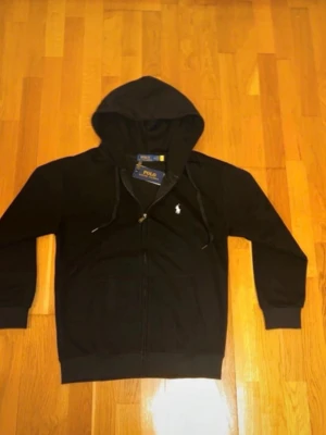 Svart hoodie från Polo Ralph Lauren - Säljer en svart hoodie från Polo Ralph Lauren med dragkedja framtill och klassisk huva. Tröjan har två fickor, ribbade muddar och den ikoniska vita Polo-loggan broderad på bröstet. Tillverkad i mjukt material för en skön och avslappnad look.