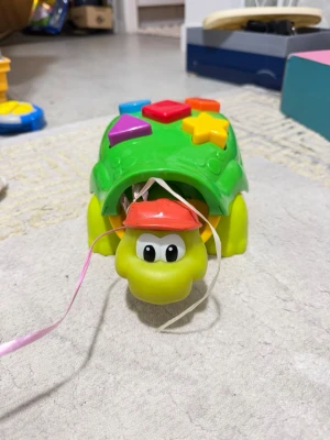 Grön sköldpaddsleksak med former - Säljer en söt grön sköldpaddsleksak i plast med färgglada former på skalet. Sköldpaddan har ett rött kepsliknande lock på huvudet och  perfekt för yngre barn som vill lära sig om olika former och färger.