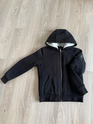 Moose knucklers hoodie - Svart hoodie med dragkedja och vit fleecefoder i huvan. Har en cool patch på vänster ärm och två fickor framtill. Ribbstickade muddar vid ärmslut och nederkant. Perfekt för en avslappnad streetstyle-look. Upplevs lite solblekt. Junior storlek XL.
