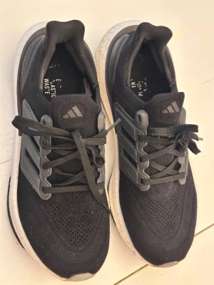 Adidas Ultraboost svarta sneakers som ny  - Svarta Adidas Ultraboost sneakers med stickad ovandel, gråa detaljer och klassiska tre ränder på sidan. Skorna har en vit, dämpad sula och snörning framtill. Tillverkade i textil och syntetmaterial, perfekt för dig som gillar sportig och modern stil.