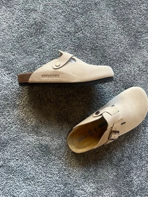  Birkenstock Boston Sand/Taube Storlek 38 - Säljer ett par snygga beige tofflor från Birkenstock i mjuk mocka. Modellen har öppen häl, justerbart spänne och präglad logga på sidan. Sulan är ergonomiskt utformad för extra komfort. Perfekta för dig som gillar stilrena och bekväma skor.