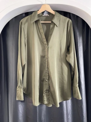 Olivgrön sidenliknande skjorta - Stilren olivgrön skjorta med lång ärm och klassisk krage. Skjortan har knappar framtill och en lös passform som ger en avslappnad look. Materialet har en sidenliknande glans som känns lyxig och mjuk mot huden.