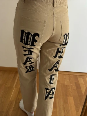 Beiga manchesterbyxor med tryck - Unika beiga manchesterbyxor med raka ben och stora svarta bokstavstryck på baksidan där det står 'CHAIN WEAR'. Byxorna har klassiska fickor bak och hällor för skärp. Perfekta för dig som vill sticka ut med streetwear-vibe.