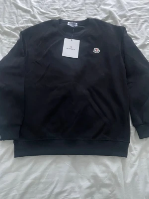 Svart sweatshirt från Moncler - Snygg svart sweatshirt från Moncler med rund halsringning och Moncler-logga broderad på bröstet. Tröjan har ribbade muddar vid ärmslut och nederkant. Perfekt för en clean och stilren look.