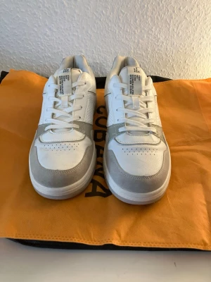 Vita och grå sneakers från Axel Arigato - Snygga sneakers från Axel Arigato i vitt och ljusgrått. Skorna har en klassisk låg siluett med ovandel i läder och mocka, perforerade detaljer på sidan och tåpartiet samt vita snören. Perfekta för en clean och stilren look.