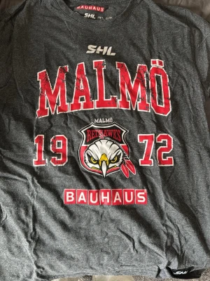 Malmö Redhawks SHL t-shirt grå - Cool grå t-shirt med stort Malmö Redhawks-tryck, röda och vita detaljer samt SHL-logga. Framsidan har årtalet 1972 och en stor örn i mitten. Perfekt för hockeyfans som vill visa sitt stöd för Malmö Redhawks.