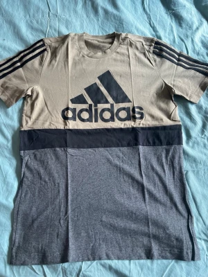 Beige och grå Adidas t-shirt - Snygg t-shirt från Adidas i beige och grå med svart logga framtill och svarta ränder på ärmarna. T-shirten har en bred svart rand över bröstet och är tillverkad i mjuk bomull. Perfekt för en sportig och avslappnad stil.
