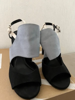 Zara Svarta och ljusgrå pumps med klack - Snygga pumps med höga klackar i svart mocka och ljusgrå mockadetalj framtill. Skorna har öppen tå och justerbar rem runt ankeln i vit. (Passar till storlek 36-37) Perfekta för dig som vill sticka ut med en unik design.