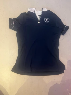 Marinblå kortärmad skjorta från Horze - Snygg marinblå kortärmad skjorta från Horka  med vit krage och dragkedja framtill. Skjortan har ett broderat märke på bröstet och är tillverkad i ett stretchigt syntetmaterial som passar perfekt till ridning. Andvänt inte jätte mycket men ändå ganska mycket men är bra skick 