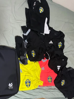 Svenska landslaget träningskläder Adidas - Säljer ett set med svenska landslagets träningskläder från Adidas. Inkluderar svarta, gula och röda tröjor med landslagets emblem, shorts och strumpor. Perfekt för fotbollsträning eller match. Väskan från b+d ingår också.