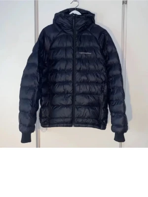 Svart dunjacka med huva Peak Performance - Snygg svart dunjacka från Peak Performance med huva och dragkedja framtill. Jackan har quiltad design, två sidofickor med dragkedja och ribbade muddar vid ärmsluten. Perfekt för kalla vinterdagar och riktigt skön att bära. Priset står inte skrivit i sten! Använd under en vinter