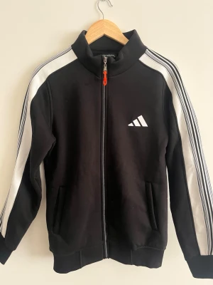 Svart Adidas ziphoodie  - Svart hoodie från Adidas. Klassisk Adidas-logga på bröstet och två fickor framtill. Tillverkad i mjukt material, perfekt för en sportig och avslappnad stil. 