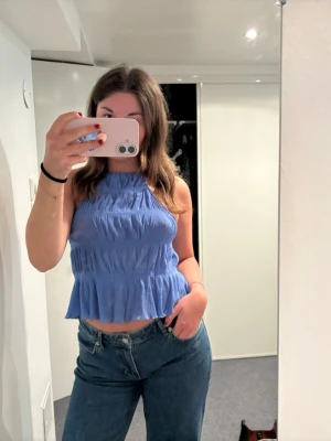 Blå ärmlös linnetopp med volang - Snygg blå linnetopp med hög hals och volangkant nedtill. Toppen är croppad och har en smockad struktur som ger en avslappnad men trendig look. Perfekt till jeans eller kjol för en fräsch stil.